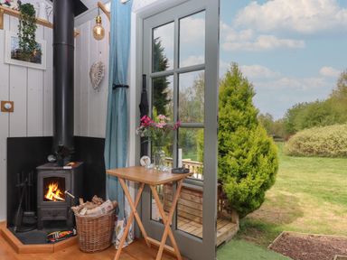 A corner of a room with a wood burning stove and a small wooden table with flowers next to a glass door opening to a garden at Burr 1 Shepherds Hut in Barnstaple