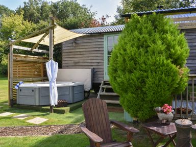 A garden with a wooden chair and table a hot tub under a canopy next to a wooden building at Burr 1 Shepherds Hut in Barnstaple