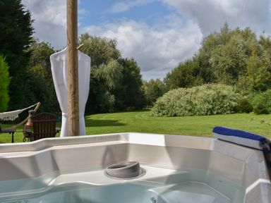 An outdoor hot tub with water and a towel on the edge overlooking a grassy yard with trees in the background at Burr 1 Shepherds Hut in Barnstaple