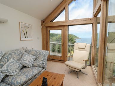 A seating area with a floral patterned sofa and a beige chair next to large wooden framed windows overlooking a sea view at Pen y Graig in Aberporth