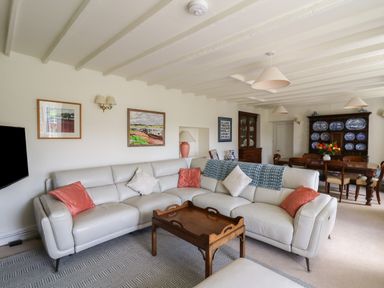 A living room with a white sectional sofa red and white cushions wooden coffee table paintings on the wall and a dining table with chairs and a cabinet with blue plates at Pen y Graig in Aberporth