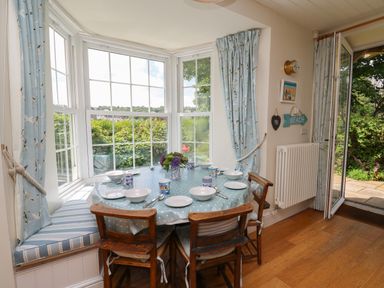 A dining area with a round table set for six by large windows and an open door at Pen y Graig in Aberporth