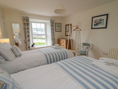 A bedroom with two single beds with striped blankets a wooden dresser a white chair a window with patterned curtains and framed pictures on the wall at Pen y Graig in Aberporth