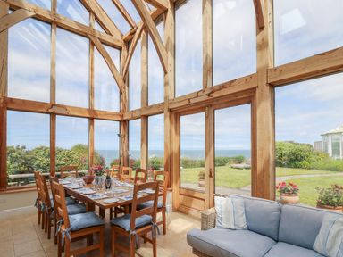 A dining area with a wooden table and chairs with cushions near large glass windows and doors showing a garden and sea at Pen y Graig Aberporth