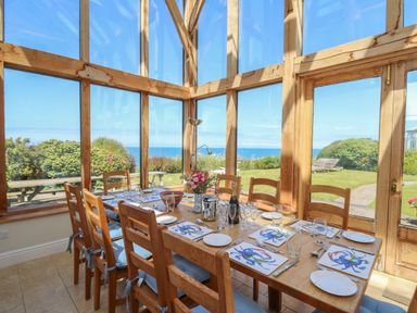 A dining area with a wooden table and chairs set with plates glasses and crab-themed placemats in a room with large windows overlooking a garden and ocean at Pen y Graig in Aberporth