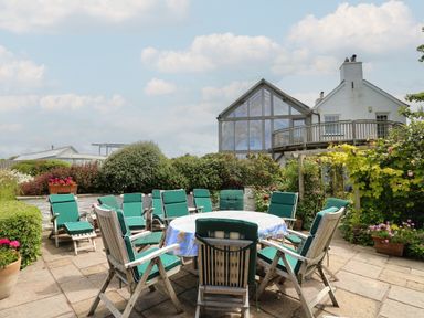 An outdoor patio with a round table covered by a tablecloth surrounded by wooden chairs with green cushions and bushes and flowers near a white house with a glass extension at Pen y Graig in Aberporth