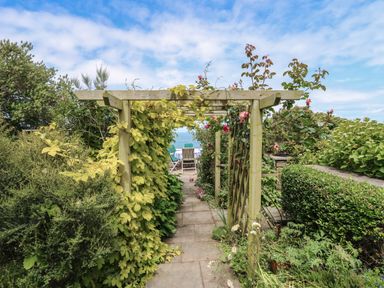 A garden walkway with a wooden pergola and green plants leading to outdoor chairs and a table at Pen y Graig in Aberporth