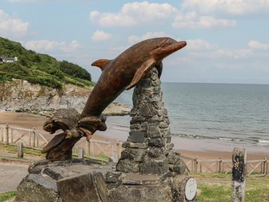 A wooden dolphin sculpture mounted on a stone pedestal near a beach with a rocky cliff and ocean in the background at Pen y Graig in Aberporth