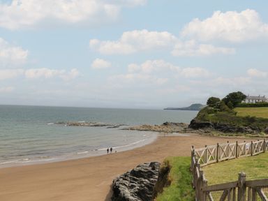 A sandy beach with rocks and a few people walking near the water by a grassy cliff with a wooden fence and a house at Pen y Graig in Aberporth