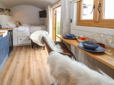 A small room with a bed a wall-mounted TV a kitchenette with blue cabinets a wooden dining counter with two plates and glasses and two chairs with white fur throws at Cariad Mawr in Gwaenysgor near Prestatyn