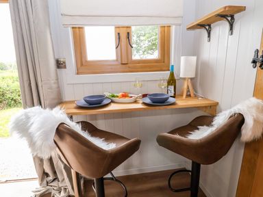A small dining area with two brown chairs with white fluffy covers at a wooden counter with plates glasses and a bottle of wine near a window at Cariad Mawr in Gwaenysgor near Prestatyn