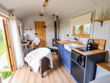 A small kitchen and dining area with blue cabinets a wood stove a brown chair with a white throw and a small table with two place settings at Cariad Mawr in Gwaenysgor near Prestatyn