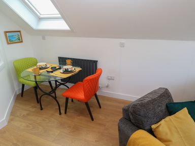 A dining area with a glass table set for two with colorful chairs and a gray sofa with cushions at Cuckoo's Rest in Penryn