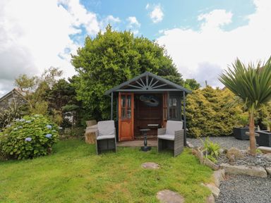 A garden with a small wooden gazebo two cushioned chairs a small round table and various plants at Cuckoo's Rest in Penryn