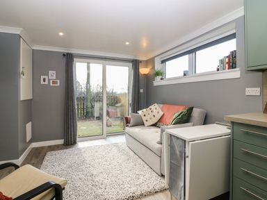 A sitting area with a sofa, cushions, a sliding glass door, curtain, a window sill with plants and books, a floor lamp, and a small table at Caldwell Cottage in Lanark