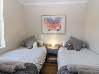 A bedroom with two single beds gray pillows a nightstand with a lamp and a framed painting on the wall at Caldwell Cottage in Lanark