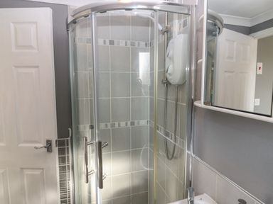 A bathroom with a glass shower enclosure a wall-mounted electric shower and a mirror above a sink at Caldwell Cottage in Lanark