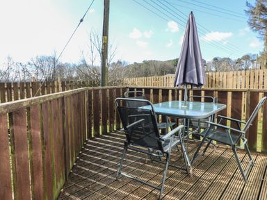 An outdoor wooden deck with a glass table shielded by a closed umbrella and four metal mesh chairs at Caldwell Cottage in Lanark