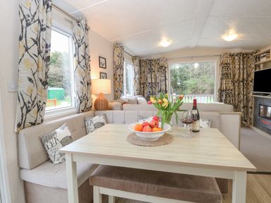 A dining area with a wooden table a bowl of fruit and tulips a bottle of wine two glasses a beige corner sofa and patterned curtains at No 3 Foxburrow Hang in Belton