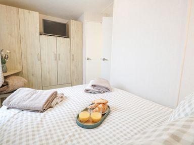 A bedroom with a bed covered in checkered bedding folded towels on the bed a tray with croissants and two glasses of orange juice wooden wardrobes with a TV on top at No 3 Foxburrow Hang in Belton