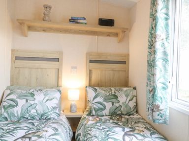 A bedroom with two single beds with tropical leaf bedding a lamp on a bedside table and a shelf with books and a game console at No 3 Foxburrow Hang in Belton