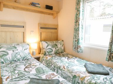 A bedroom with two single beds with patterned bedding a bedside table with a lamp a wooden shelf with books and a device and a window with patterned curtains at No 3 Foxburrow Hang in Belton