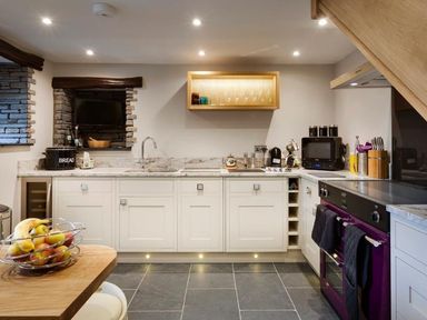 A kitchen with white cabinets a purple oven a fruit bowl on a wooden table and a mounted wooden shelf at The Cosy Peacock in Troutbeck
