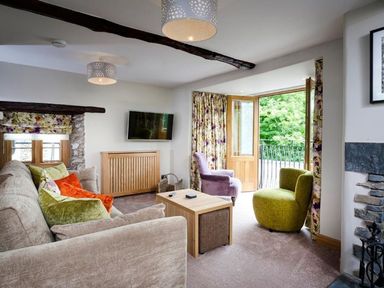 A living room with a beige sofa green and orange cushions purple and green chairs wooden table and floral curtains at The Cosy Peacock in Troutbeck