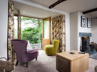 A living room with a purple armchair a green chair a wooden coffee table floral curtains and a fireplace at The Cosy Peacock in Troutbeck