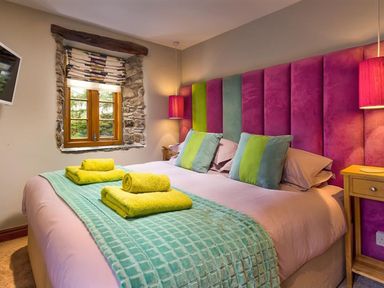 A bedroom with a bed with colorful headboard two bedside tables window with stone frame and folded towels on bed at The Cosy Peacock in Troutbeck