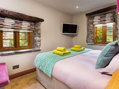 A bedroom with a bed with folded towels and pillows and two windows with patterned blinds at The Cosy Peacock in Troutbeck