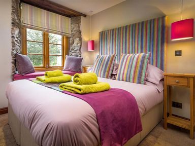 A bedroom with a double bed colorful striped headboard and pillows green towels on the bed wooden side table and window seat at The Cosy Peacock in Troutbeck