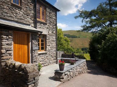 A stone house with wooden door and windows with plants on the porch and hills in the background at The Cosy Peacock in Troutbeck