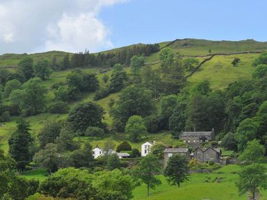 Houses surrounded by trees on a hillside with a blue sky at The Cosy Peacock in Troutbeck