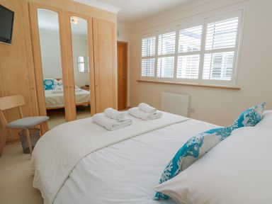 A bedroom with a bed with white bedding and blue patterned cushions a wooden chair a wardrobe with mirrored doors and white window shutters at Mariners Watch in Carbis Bay
