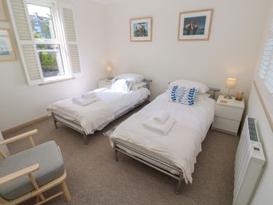 A bedroom with two single beds each with white bedding and towels bedside tables with lamps a window with shutters and a chair at Mariners Watch in Carbis Bay