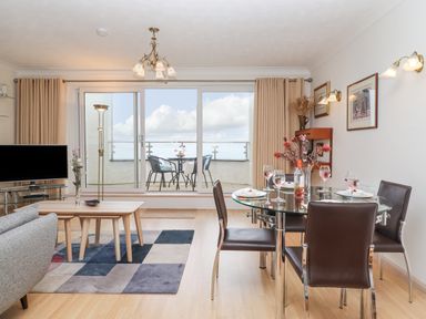 A dining area with a glass table set with glasses and plates next to a living area with a TV and coffee tables near a balcony with outdoor chairs and a table at Mariners Watch in Carbis Bay