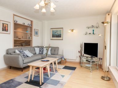 A living room with a gray sectional sofa and patterned cushions two nested wooden coffee tables on a multicolored rug a TV on a glass stand and wall shelves with decor at Mariners Watch in Carbis Bay