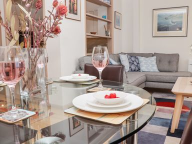 A dining table with plates, bowls of raspberries, wine glasses with pink liquid, and a vase with dried flowers in a living room with a gray sofa and cushions at Mariners Watch in Carbis Bay