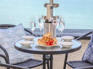 A round glass table with croissants strawberries four bowls four glasses of water and a bottle in an ice bucket with flowers on a balcony with two chairs and cushions at Mariners Watch in Carbis Bay