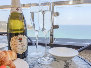 A bottle of Prosecco with two filled glasses on a glass table with a croissant and a white dish on a balcony overlooking the sea at Mariners Watch in Carbis Bay