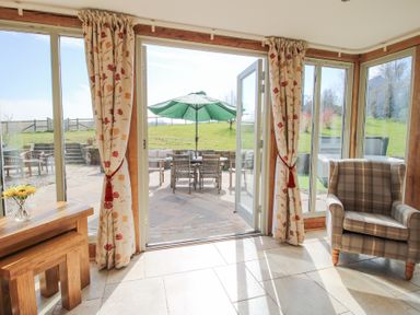 A sunroom with floral curtains a wooden table a plaid armchair and glass doors opening to a patio with outdoor furniture and a green umbrella at Cuckoo Pen Cottage Eastham near Tenbury Wells