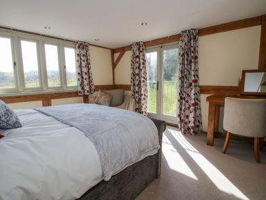 A bedroom with a bed a chair a wooden desk with a mirror and floral curtains at Cuckoo Pen Cottage in Eastham near Tenbury Wells