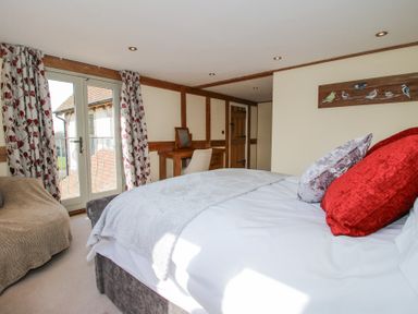 A bedroom with a bed red and silver pillows a chair and a desk near French doors with floral curtains at Cuckoo Pen Cottage in Eastham near Tenbury Wells