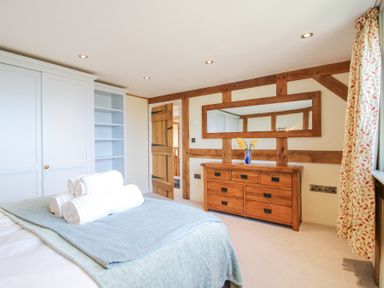 A bedroom with a bed, folded towels, wooden dresser with a vase, large mirror, wooden door, built-in white wardrobe, and floral curtains at Cuckoo Pen Cottage in Eastham near Tenbury Wells