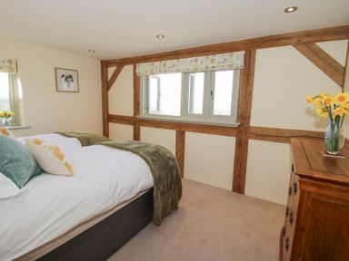A bedroom with a bed cushions and a blanket a wooden dresser with a vase of flowers and windows with floral blinds at Cuckoo Pen Cottage in Eastham near Tenbury Wells