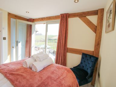 A bedroom with a bed covered by a pink blanket and rolled white towels on it next to a blue chair and open doors leading to a balcony with a view of green fields at Cuckoo Pen Cottage in Eastham near Tenbury Wells