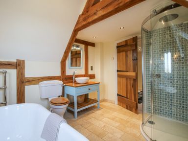 A bathroom with a bathtub a toilet a sink on a blue vanity a wooden door and a glass shower with patterned tiles at Cuckoo Pen Cottage in Eastham near Tenbury Wells