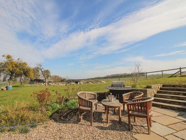 An outdoor seating area with wooden chairs, a table, and a barbecue grill in a garden with grass and sheep in the background at Cuckoo Pen Cottage in Eastham near Tenbury Wells
