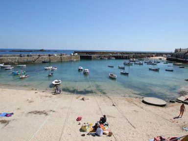 A beach with people sitting on sand and boats anchored in a harbor at The Net Loft in Mousehole